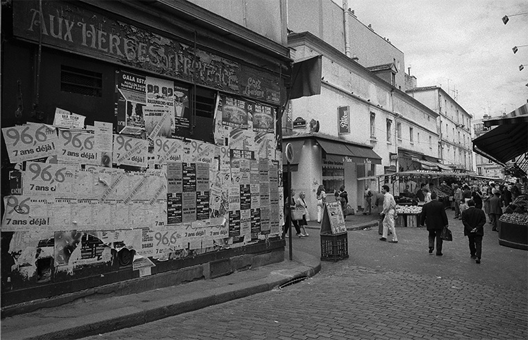 Pendant l'été 1987 à Paris, un commerce voit ses façades envahies par les affiches du moment, à proximité du marché.